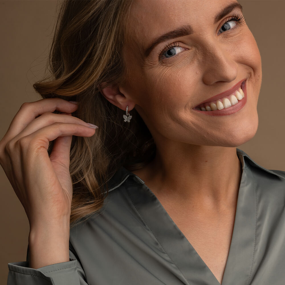 Et par sølvfargede øredobber med små glitrende stener, formet som sommerfugler som henger fra sirkulære ørekroker dekorert med matchende stener.