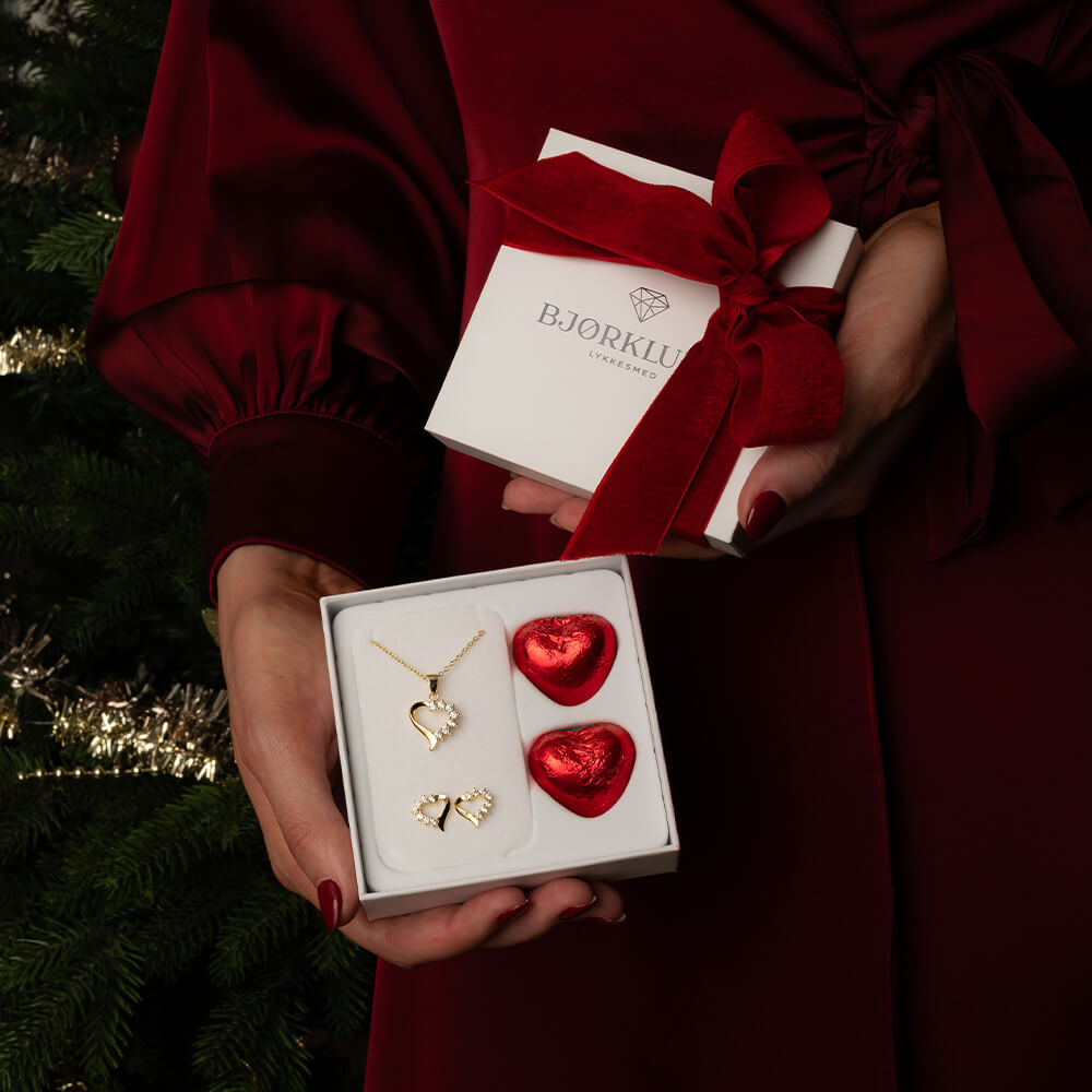 Gullfarget smykkesett med halssmykke, ring og ørestikker i asymmetrisk hjerte-design dekorert med gjennomsiktige steiner mot hvit bakgrunn