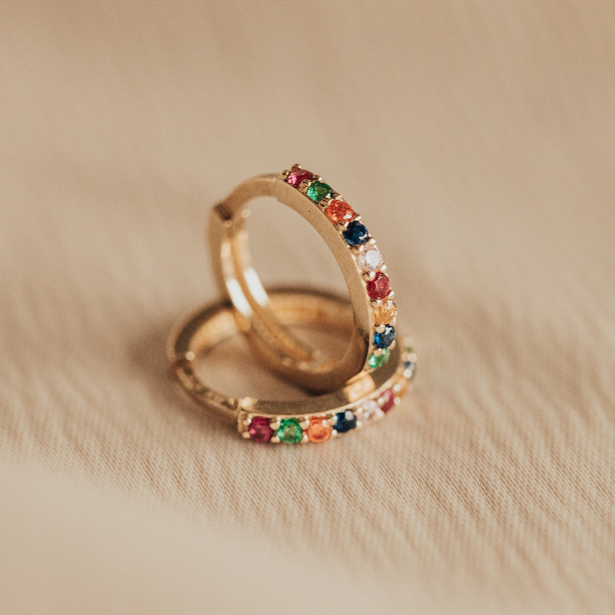 To små gullfargede ringformede øredobber med hengslet lukking, dekorert med små steiner i ulike farger, fotografert mot hvit bakgrunn.