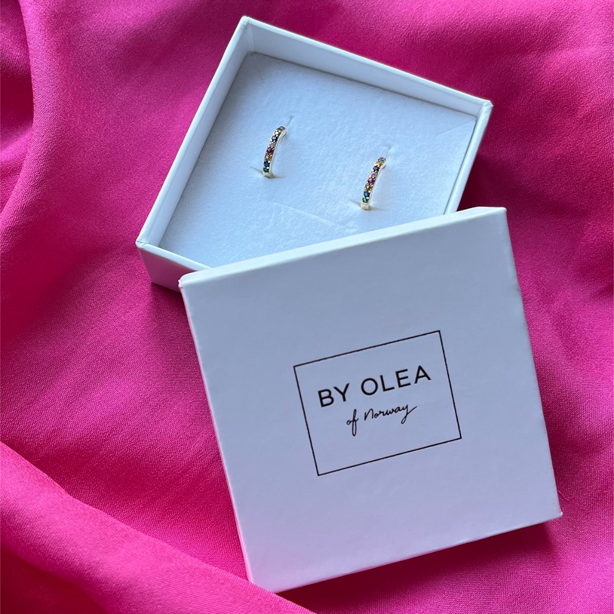 To små gullfargede ringformede øredobber med hengslet lukking, dekorert med små steiner i ulike farger, fotografert mot hvit bakgrunn.