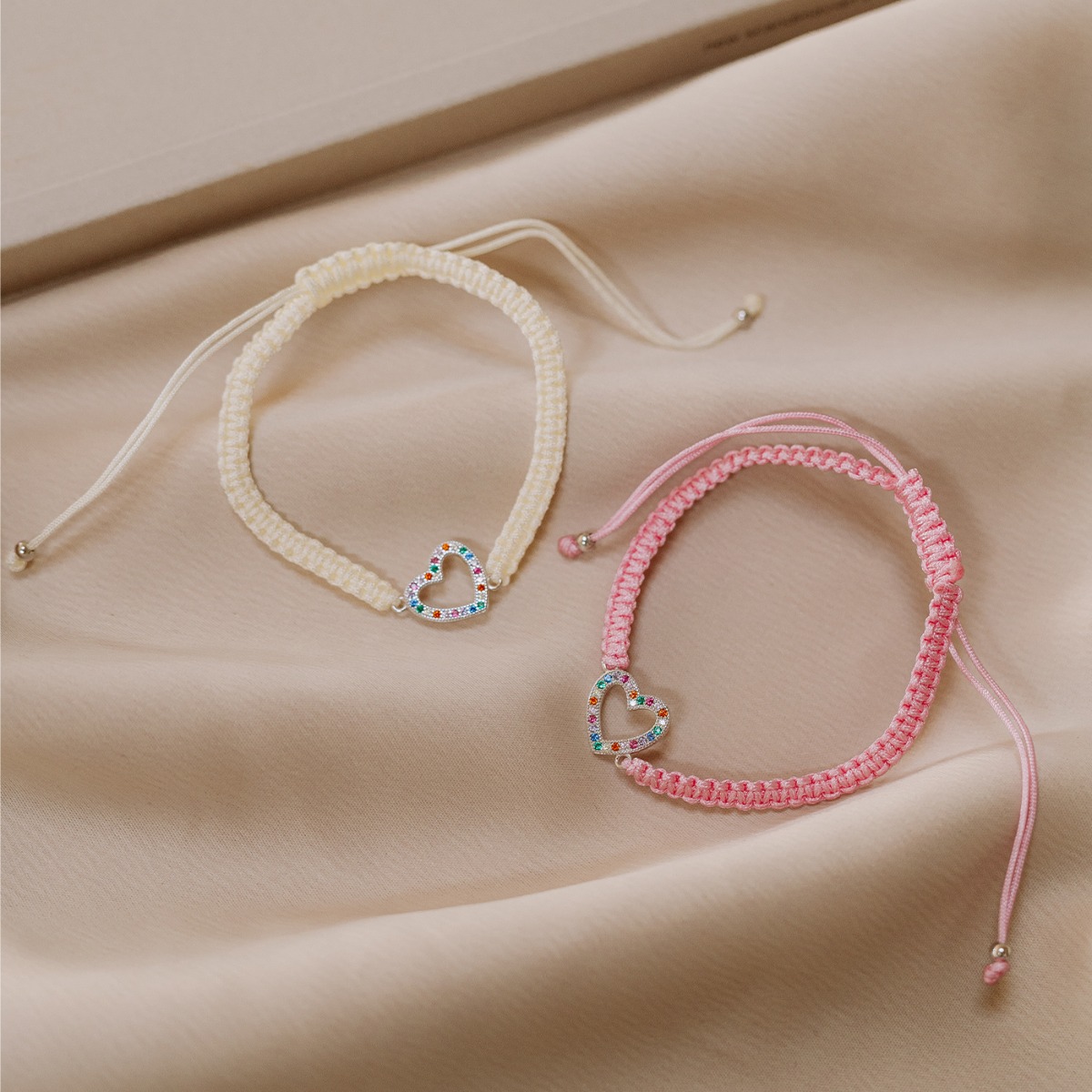 Et Frelsesarmeen vennskapsarmbånd i rosa med fargerikt hjertesmykke på hvit bakgrunn og rød Frelsesarme-logo øverst til venstre.