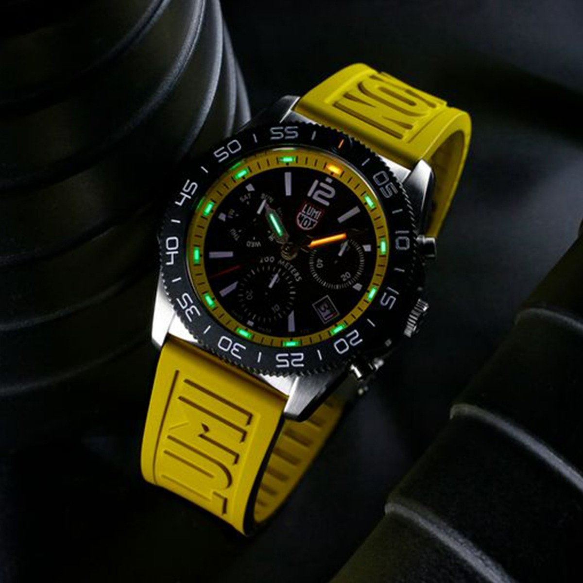 Sølvfarget herreklokke med sort og gul urskive, kronografer og lenkearmbånd, vist med ekstra gult gummireim ved siden av.