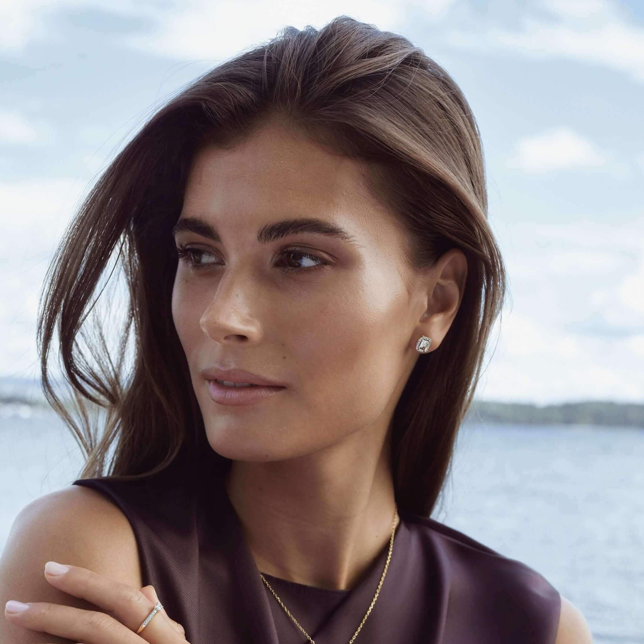 Nærbilde av kvinne med brunt hår ves sjøen. Hun har på seg brun topp og diamantsmykker