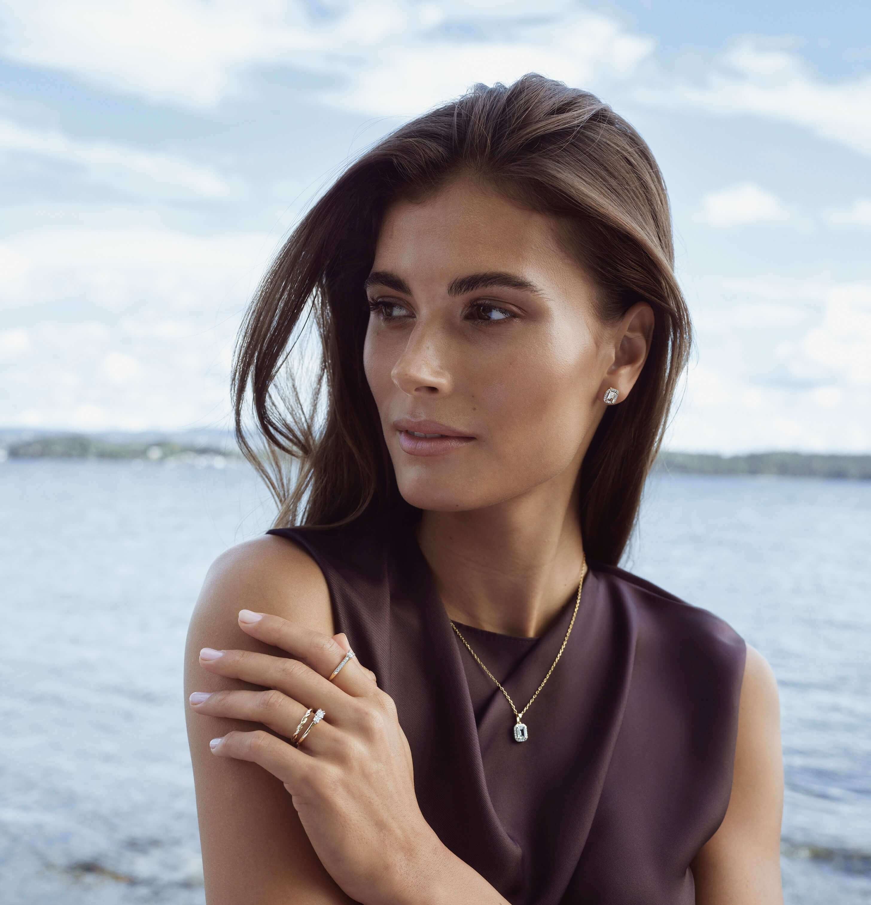 Se våre House of Piene-smykker En kvinne med mørkeblondt hår og en lys kjole, ser ut av vinduet mens hun står i mellom gardinene.