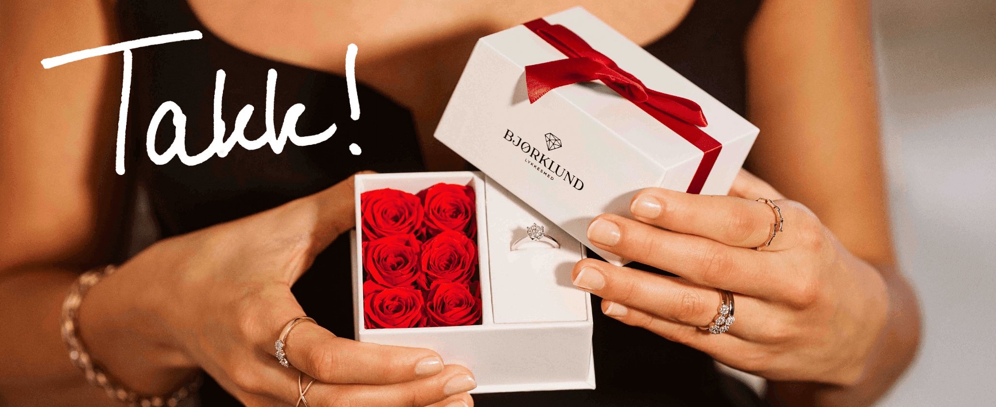 Nærbilde av en kvinne som åpner en hvis gaveeske fra Bjørklund med røde roser og en diamantring i hvitt gull. Kvinnen har mange diamantringer på hånden. Over bildet står det en tekst med hvit skrift som sier "takk!"