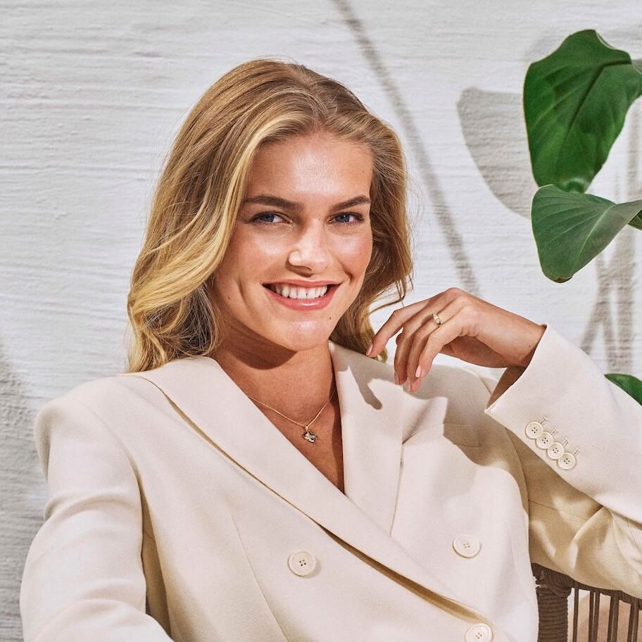 Smilende kvinne med blondt hår og kremfarget dress viser frem gullsmykke og ring.