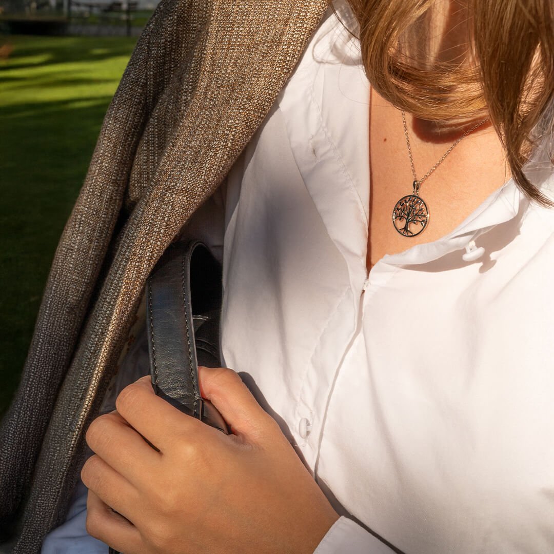 Kvinne med blondt hår bærer hvit skjorte, grå blazer og halskjede med livets tre-anheng. Hun holder en svart håndveske.