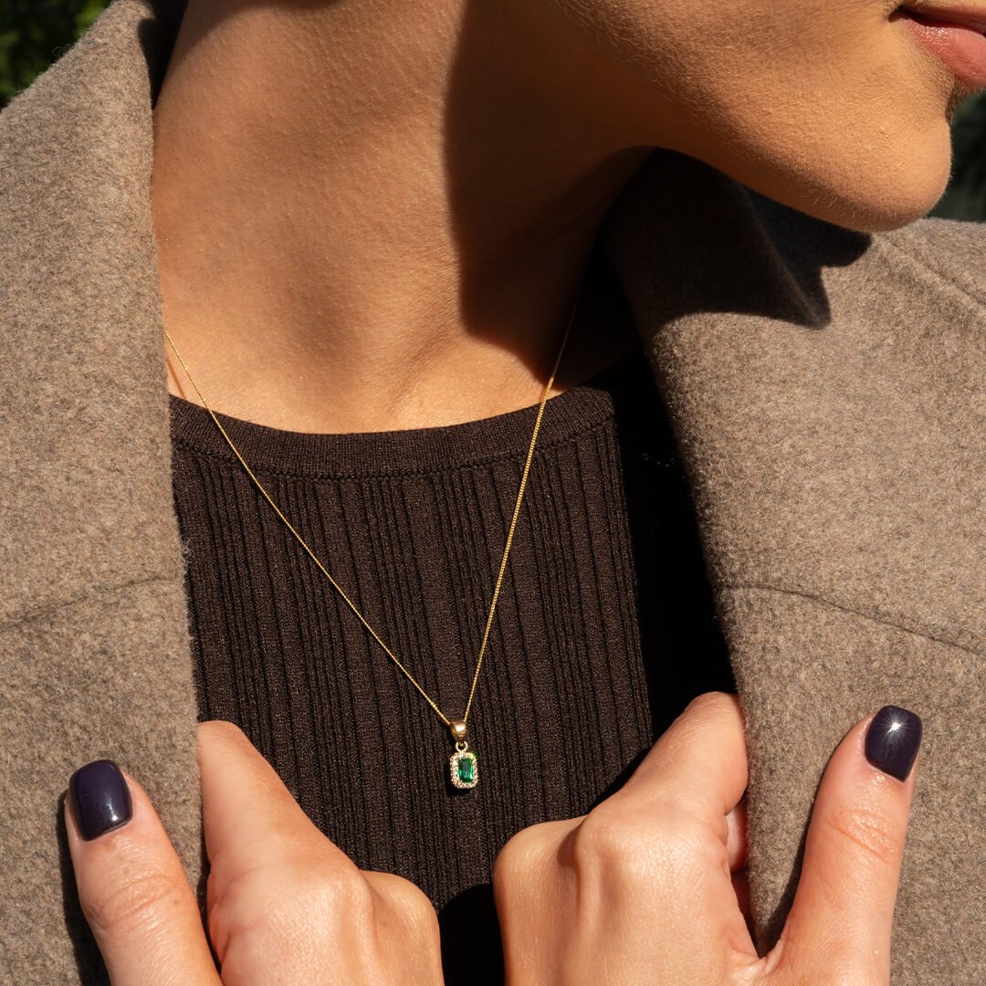 Person med mørk neglelakk på neglene holder hendene foran seg og viser et gull halskjede med grønt smaragdfarget anheng.