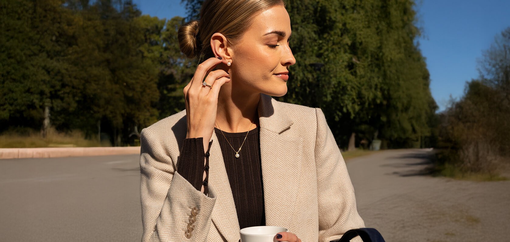 Elegant kvinne med oppsatt hår i beige fishbone-blazer og mørk topp, bærer gullring og øredobber mens hun holder hvit kaffekopp i høstlig sollys
