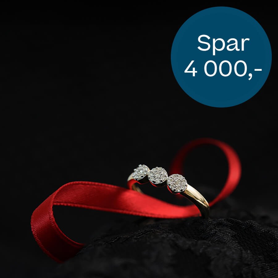 Gullring med tre små diamantklynger ligger på svart underlag med en rød silkebåndsløkke og rund blå prislapp i hjørnet.