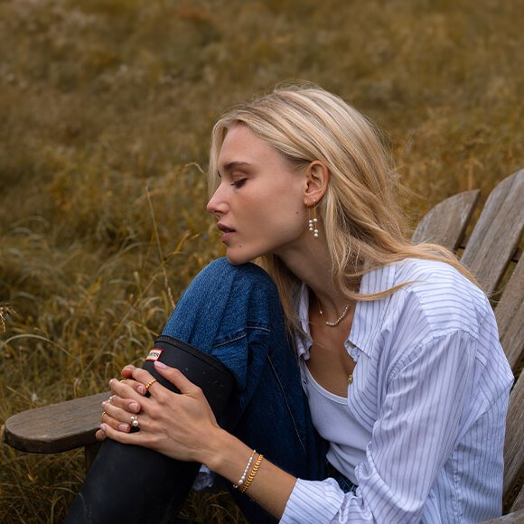 Se Ocean Flow-kolleksjonen Et par kvinnehender med seks byBiehl Ocean Flow-ringer med perler og diamanter, med fingerene i et garn som er hengt opp ute.