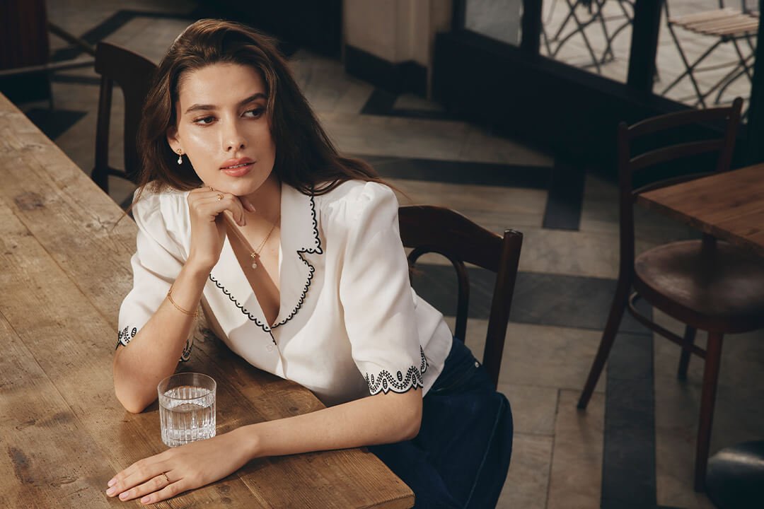 Halskjede – i gull eller sølv, med perler, symboler og plater En kvinne sitter ved et trebord på en kafé, iført en hvit bluse med svarte detaljer, gullkjede, øredobber og armbånd.