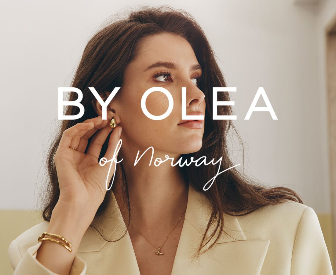 Kvinne i beige blazer berører øret sitt og viser gullfargede øredobber, halskjede, ring og armbånd.