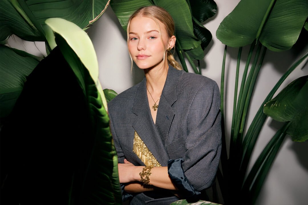 Ung kvinne med lyst hår og grå blazer står blant store grønne blader, iført gullsmykker med detaljer og strukturert design.