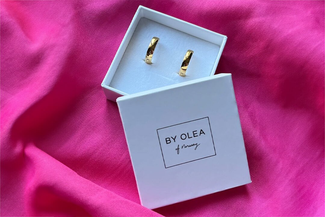 Et par brede og gullbelagte stål øreringer HEDDA i en hvit gaveeske med By Olea of Norway på lokket, ligger oppå en rosa duk.