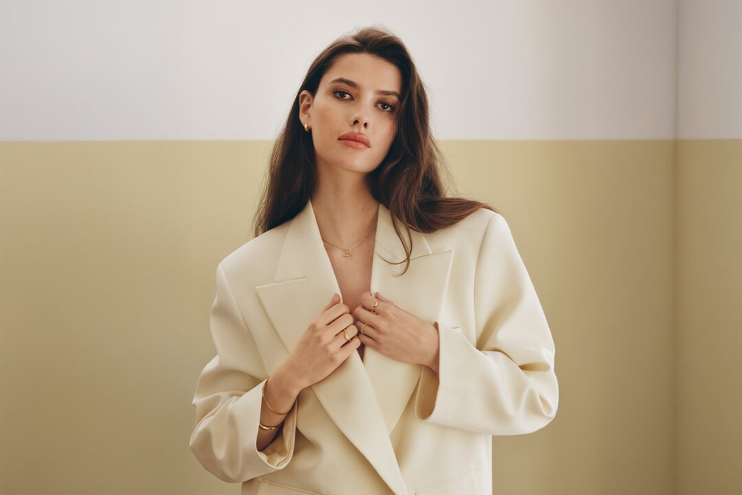 Ringer – gullfarget og med vakre detaljer og stener En kvinne med langt mørkt hår iført en oversized kremhvit blazer, stylet med flere gullsmykker, ringer og armbånd.
