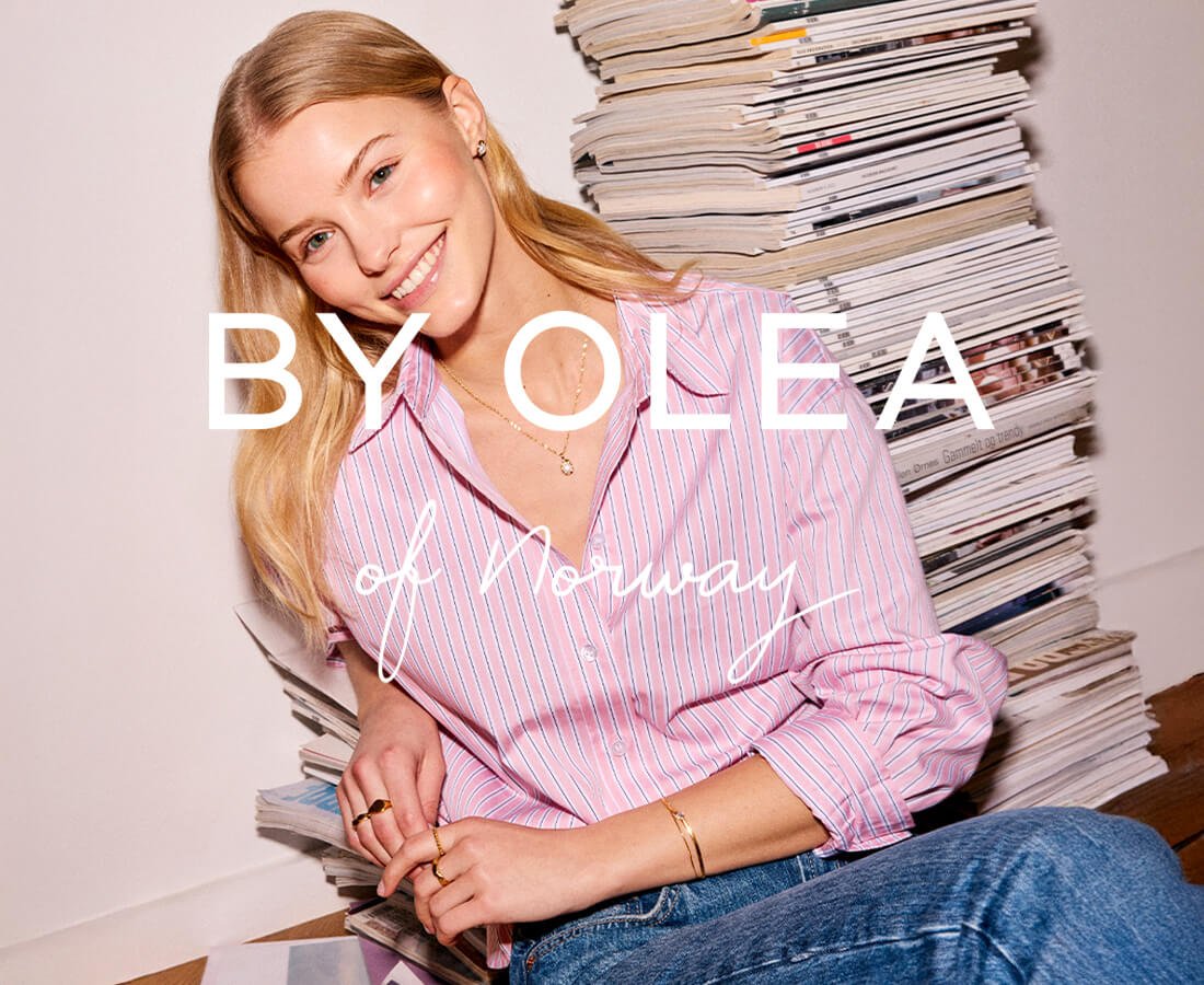 Kvinne med rosa skjorte og smykker fra By Olea sitter foran en stabel magasiner og smiler mot kamera i en uformell setting.