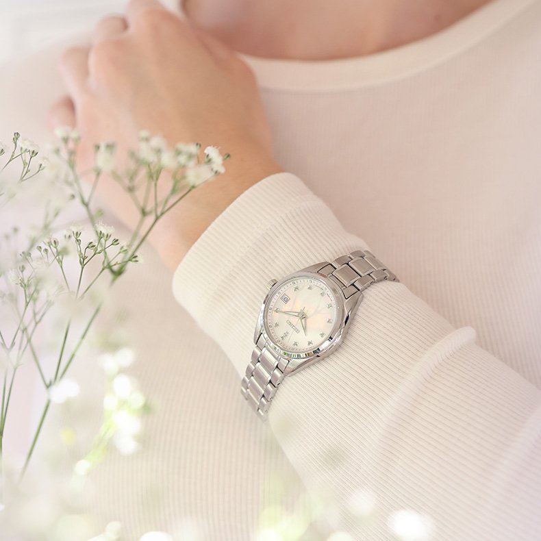 Sølvfarget dameklokke med lenke og perlemorskive på håndledd, omgitt av myke blomster i lys setting.