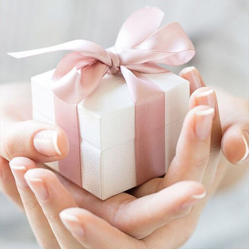 En liten, hvit gavepakke med bredt, rosa bånd knytt rundt, ligger midt i en kvinnehånd holdt av sin andre hånd, foldet.