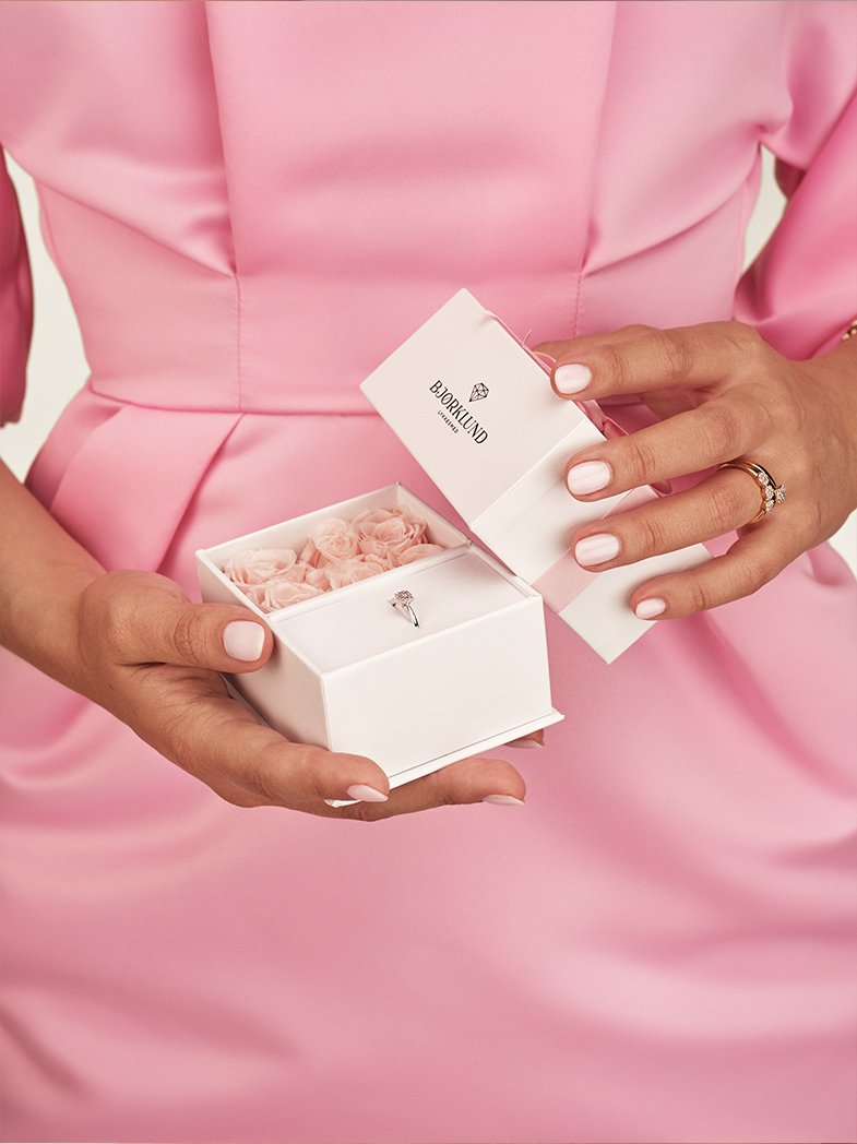 Kvinne i rosa kjole holder en Bjørklund-eske med lyserosa roser og en diamant-ring i hvitt gull som ligger i gaveesken.