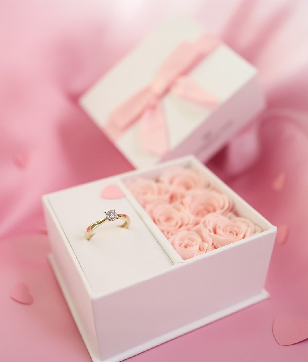Gullring med diamant ligger i hvit gaveeske med rosa roser og sløyfe mot rosa bakgrunn.