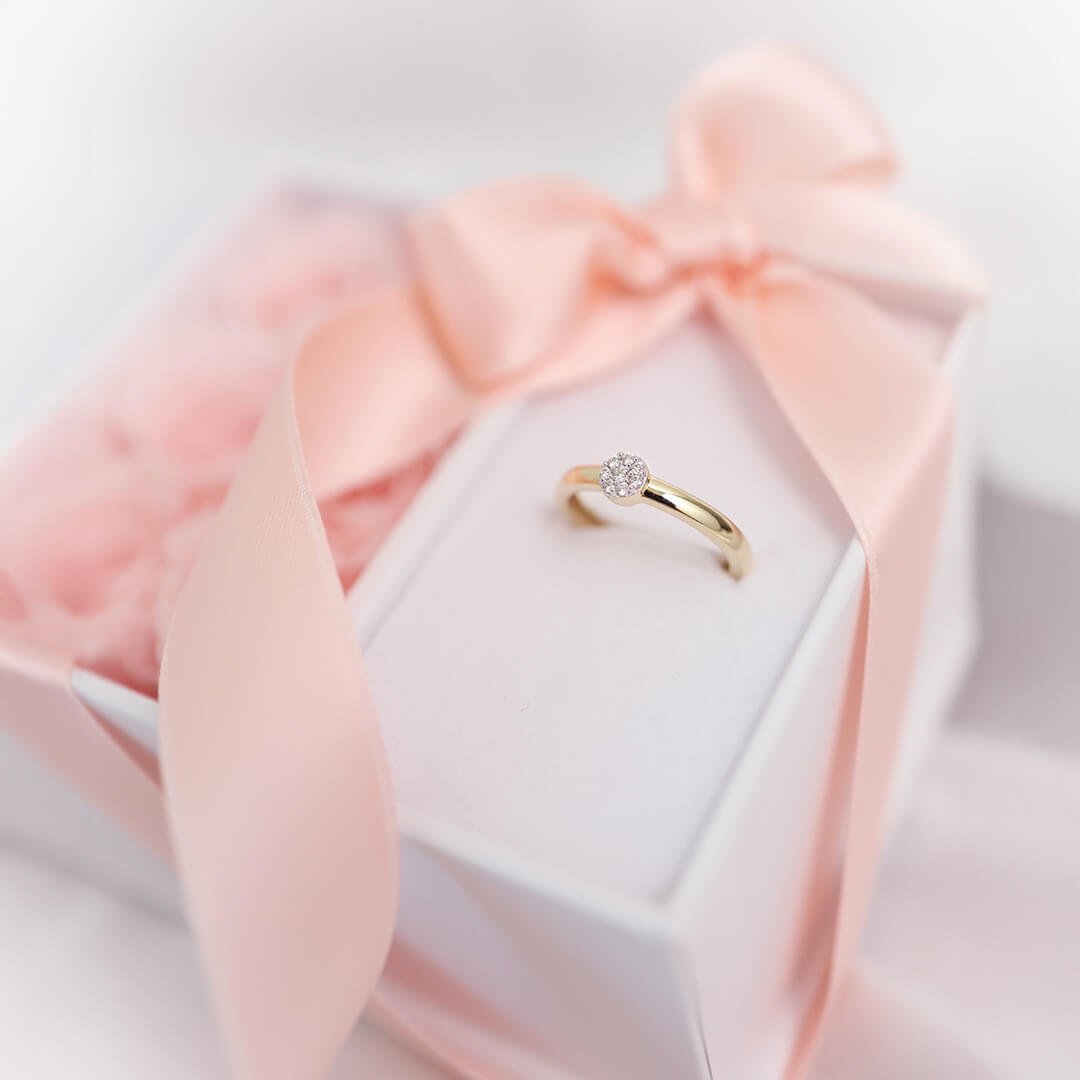 Gullring med liten diamant står på hvit gaveeske pyntet med lys rosa silkebånd.