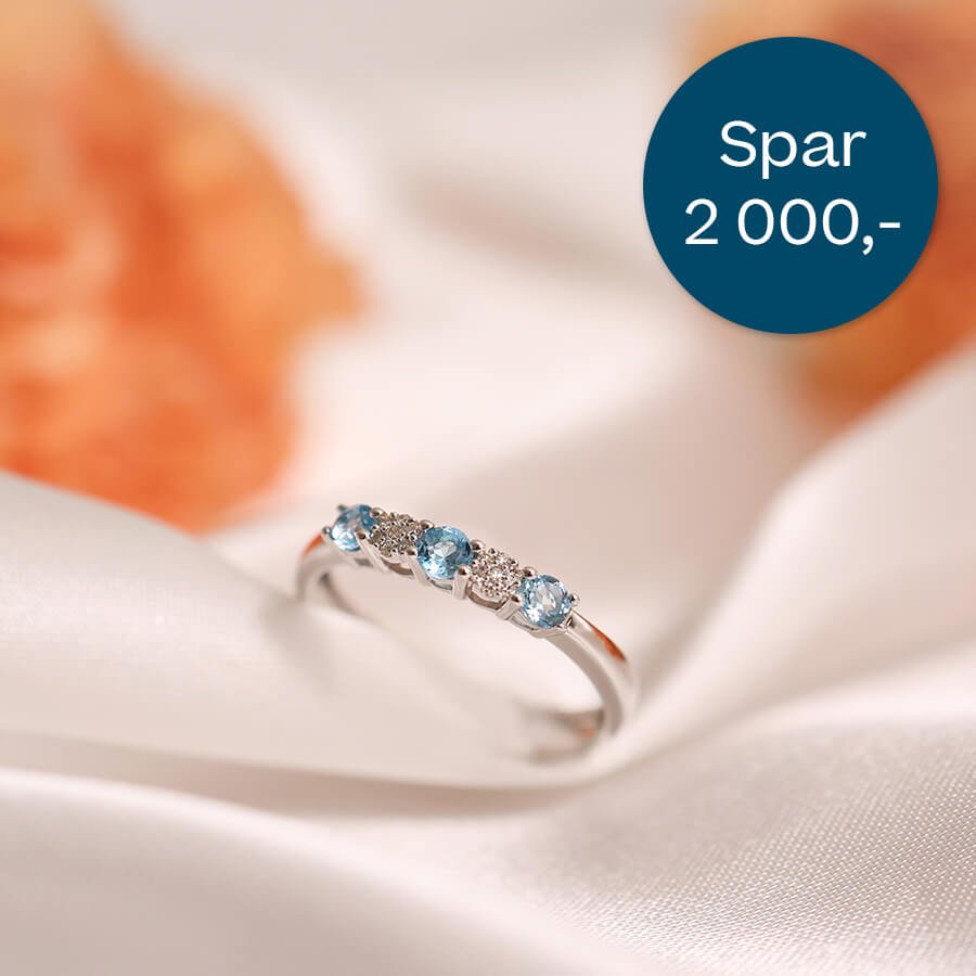 Sølvfarget ring med lyseblå steiner og små klare steiner, plassert på lys tekstilbakgrunn, med blå sirkel som viser prisavslag.