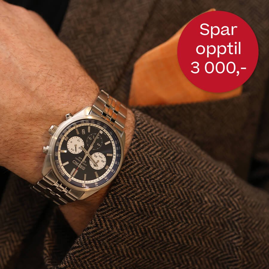 Manns håndledd med sølvfarget klokke på metallrem, iført brun dressjakke, med rød sirkel med tekst om prisavslag.