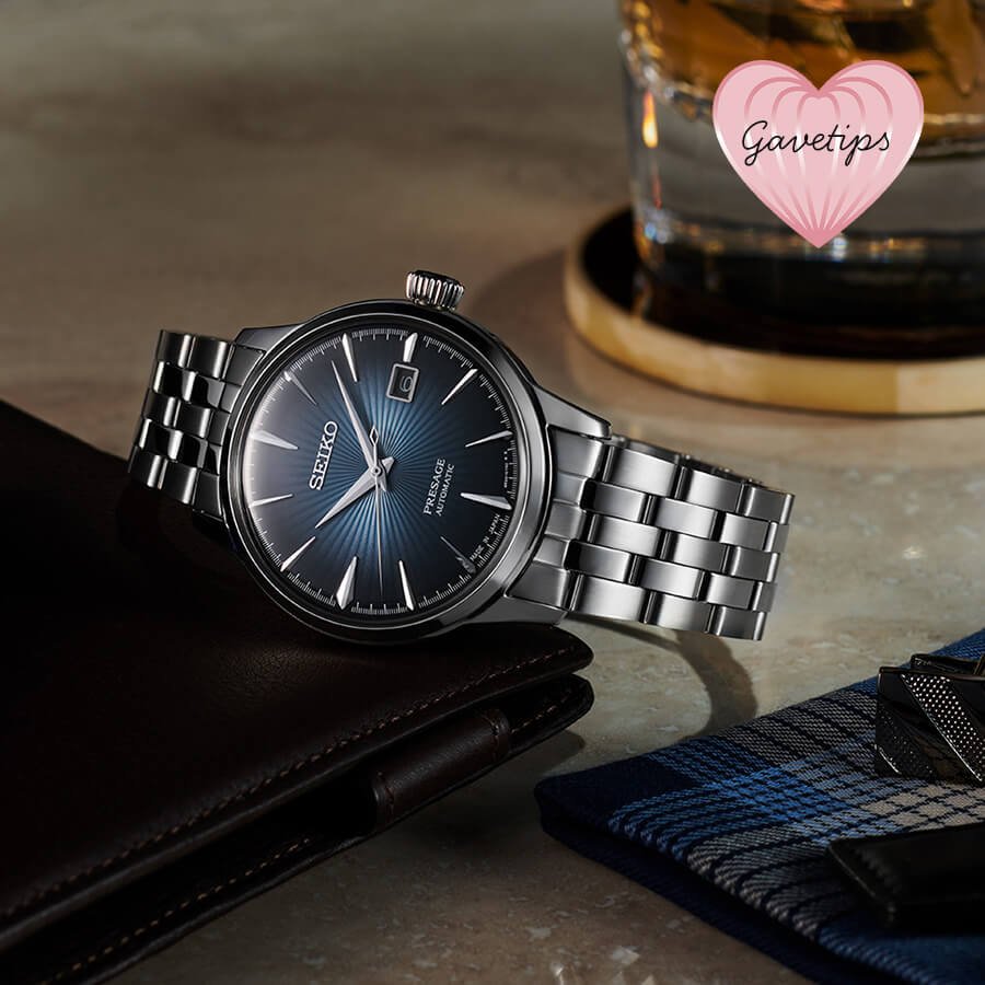 Seiko klokke med blå urskive og stållenkebånd plassert ved lommebok, slips og whiskeyglass. Rosa hjerteikon med tekst 'Gavetips'.
