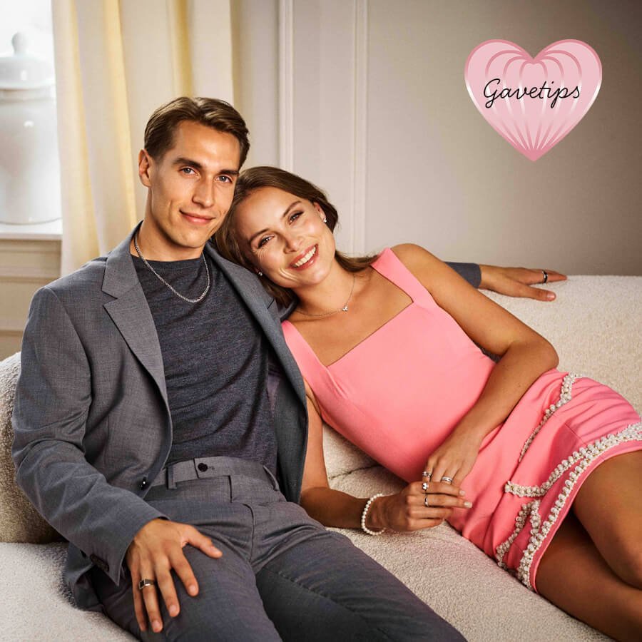 Par i elegant antrekk sitter i sofa. Kvinnen i rosa kjole bærer halskjede, ringer og armbånd. Mannen i grå dress har ring og halskjede.