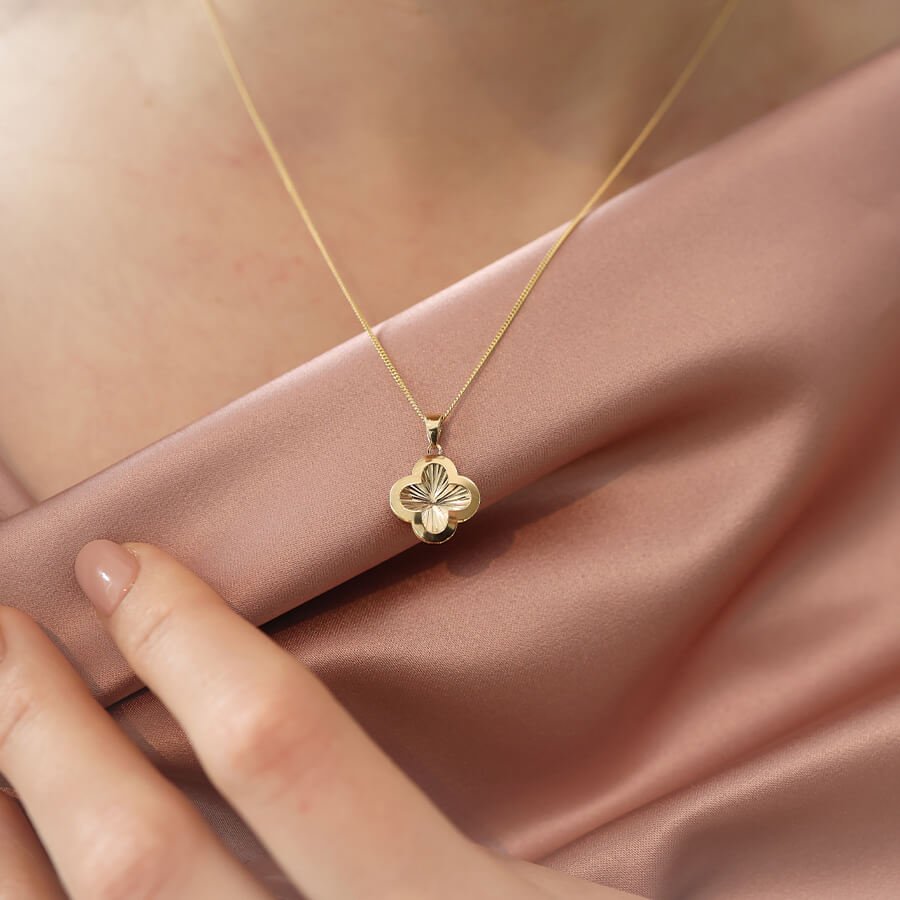 Gullkjede med et firkløverformet anheng med riller, båret over et rosa silketøy. En hånd hviler nederst i venstre hjørne.