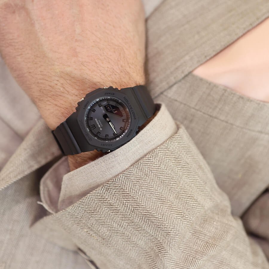 Nærbilde av en sort Casio G-Shock-klokke på et håndledd, delvis dekket av en beige tekstiljakke med strukturveving.