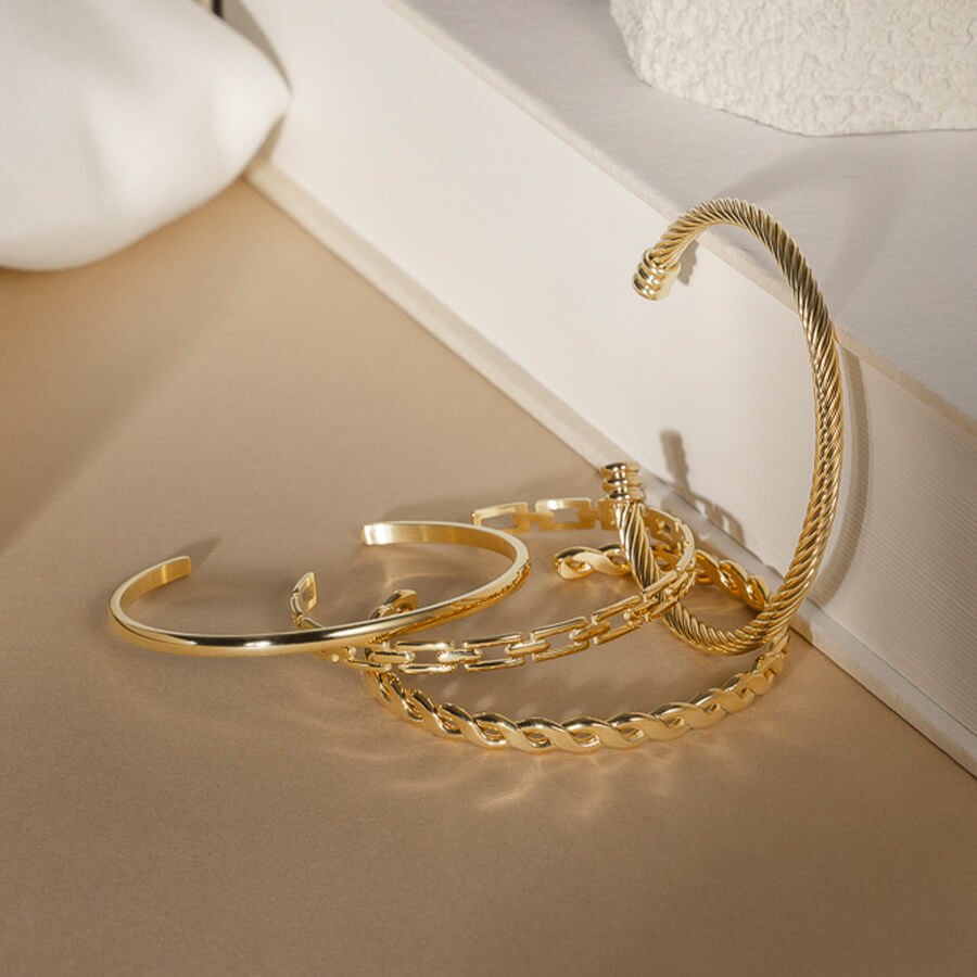 Fire gullsmykker ligger samlet mot en hvit bok og beige bakgrunn. Inkluderer en tvinnet armring og tynnere kjeder.