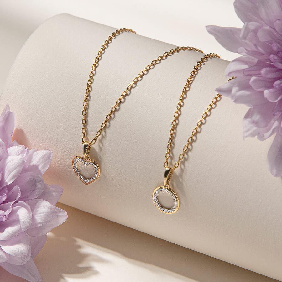 To gullkjeder med anheng i form av et hjerte og en sirkel, pyntet med glitrende steiner, drapert over en lys flate.