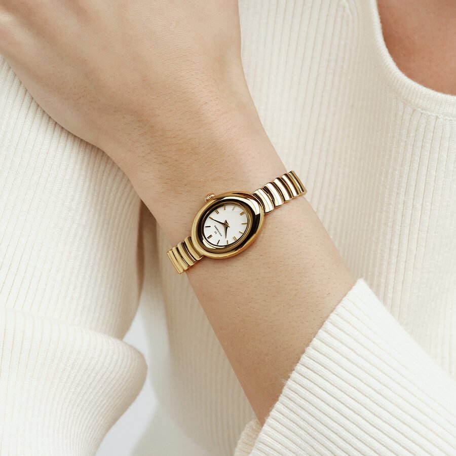 Nærbilde av en gullklokke med hvit urskive og leddrem på et håndledd. Personen har på seg en hvit, ribbestrikket genser.