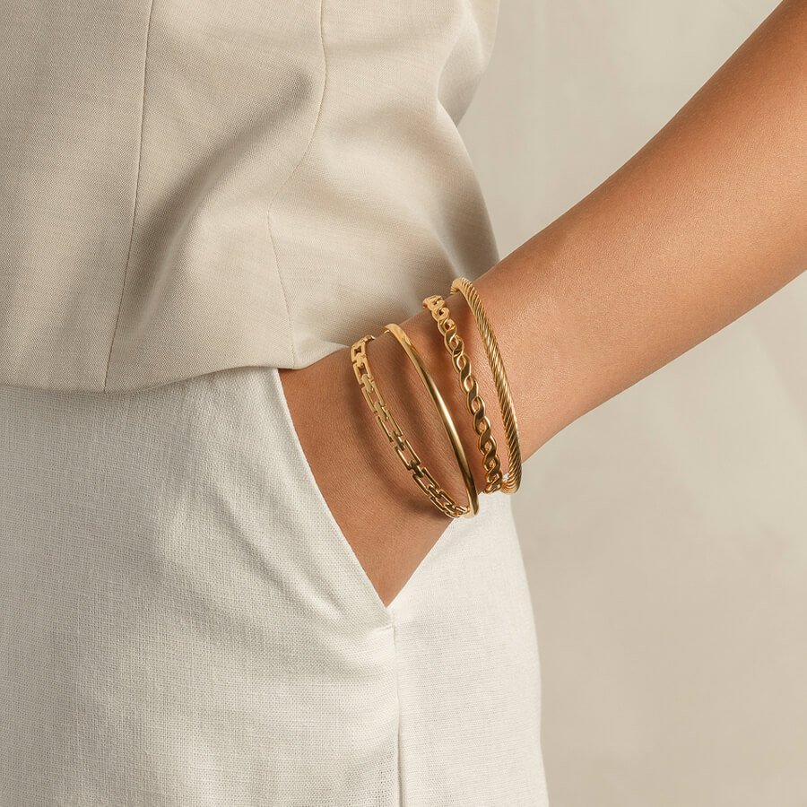 Fire gullfargede armbånd i ulike design bæres på en arm med hånden i lomma, mot nøytral bakgrunn og lyse klær.