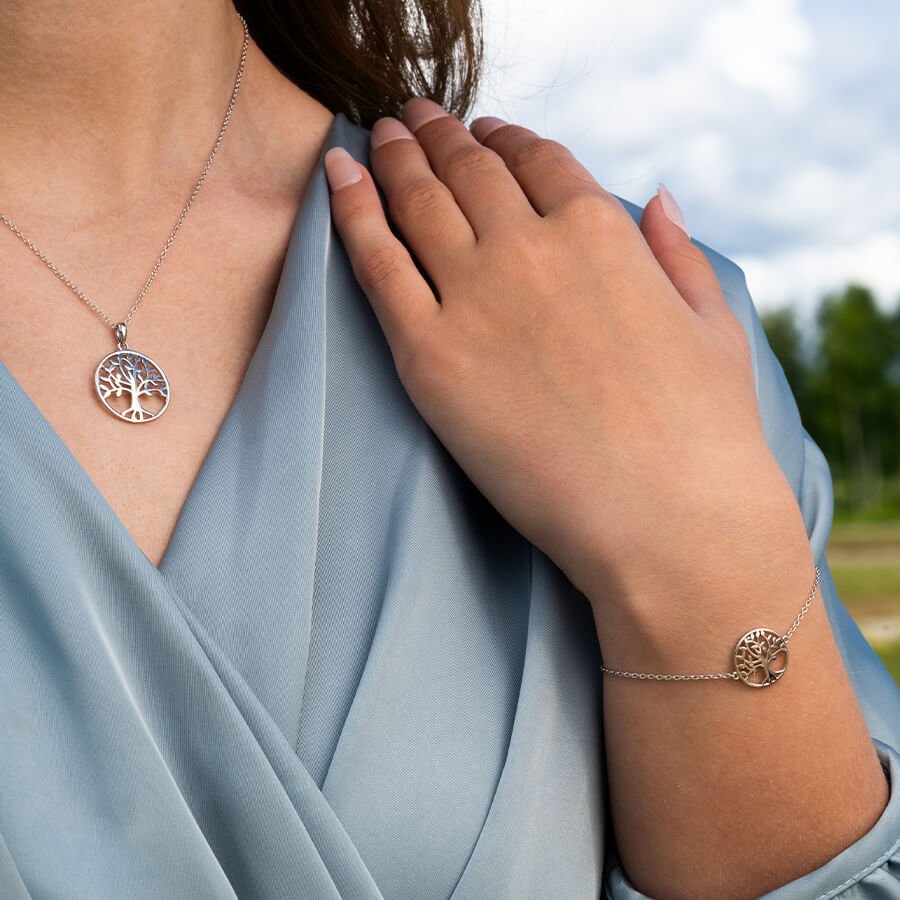 Kvinne med blå omslagskjole viser frem sølvfarget halskjede og armbånd med livets tre-symbol utendørs i dagslys.