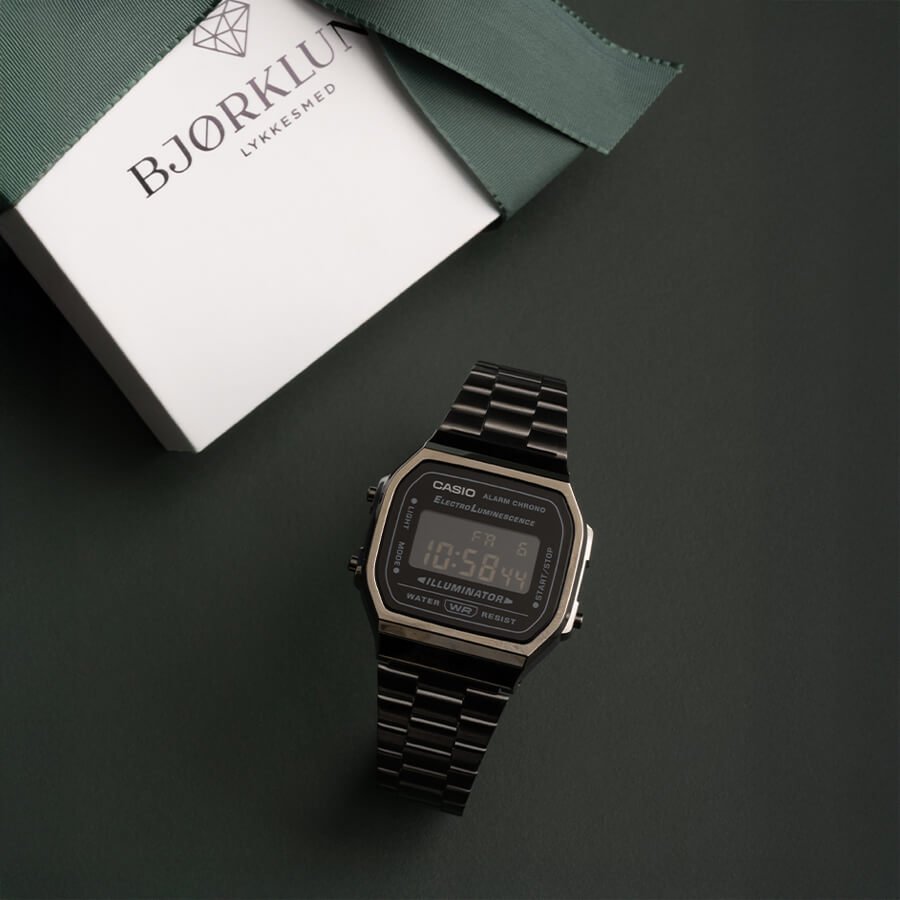 Casio digital klokke i sort metall med firkantet skjerm på mørk bakgrunn ved siden av Bjørklund-gaveeske i grønt og hvitt.