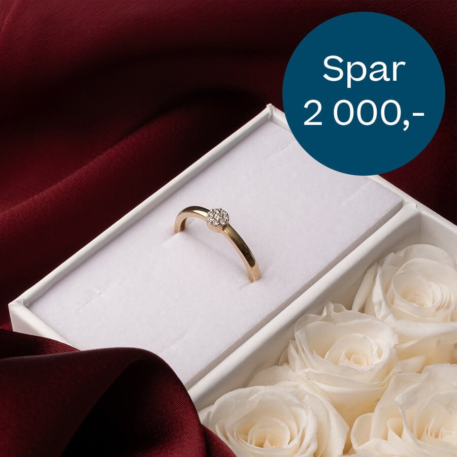 Forlovelsesring i gull med steiner i hvit gaveeske ved siden av boks med hvite roser på mørkerød bakgrunn.