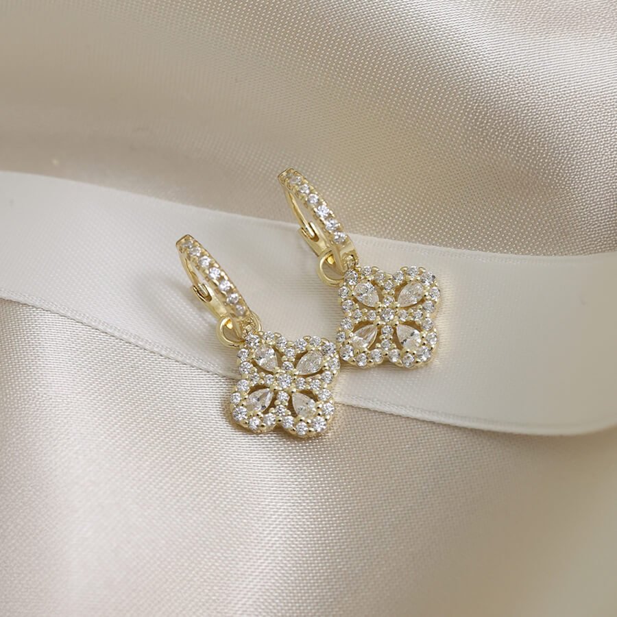 Gullfargede øredobber med blomstformet anheng besatt med små diamanter, lagt på lys sateng med dekorative strådetaljer.