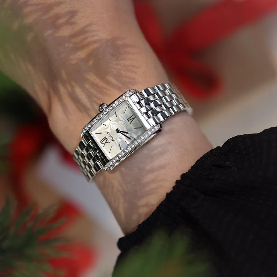 Sølvfarget rektangulær klokke med lenke på en håndledd, fotografert i nærbilde med uskarpe grønne og røde elementer i forgrunnen.