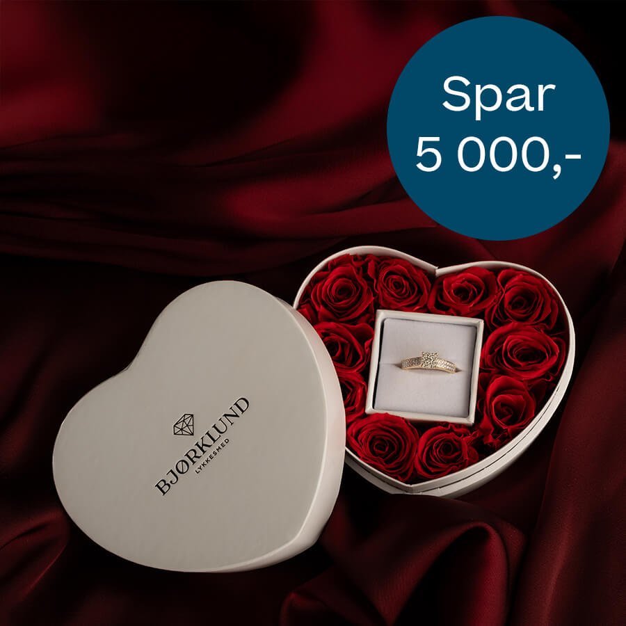 Hjerteformet eske med røde roser og en ring i midten, plassert på mørkerødt stoff ved siden av lokket med Bjørklund-logo.