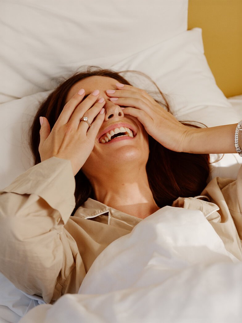 Kvinne ligger i seng og ler med hendene over øynene, iført beige skjorte, med forlovelsesring synlig på fingeren.