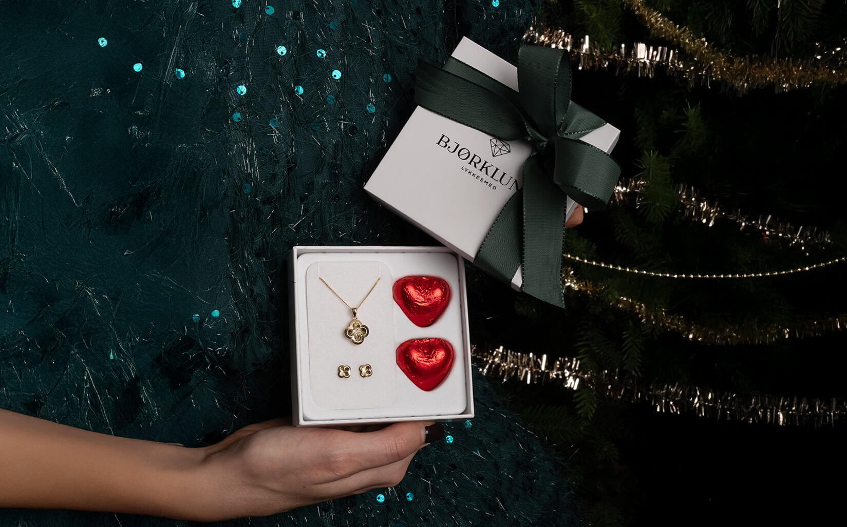 Våre klassiske gaveesker! Hender holder Bjørklund-gaveeske med gullsmykkesett og to røde hjerteformede sjokolader foran juletre