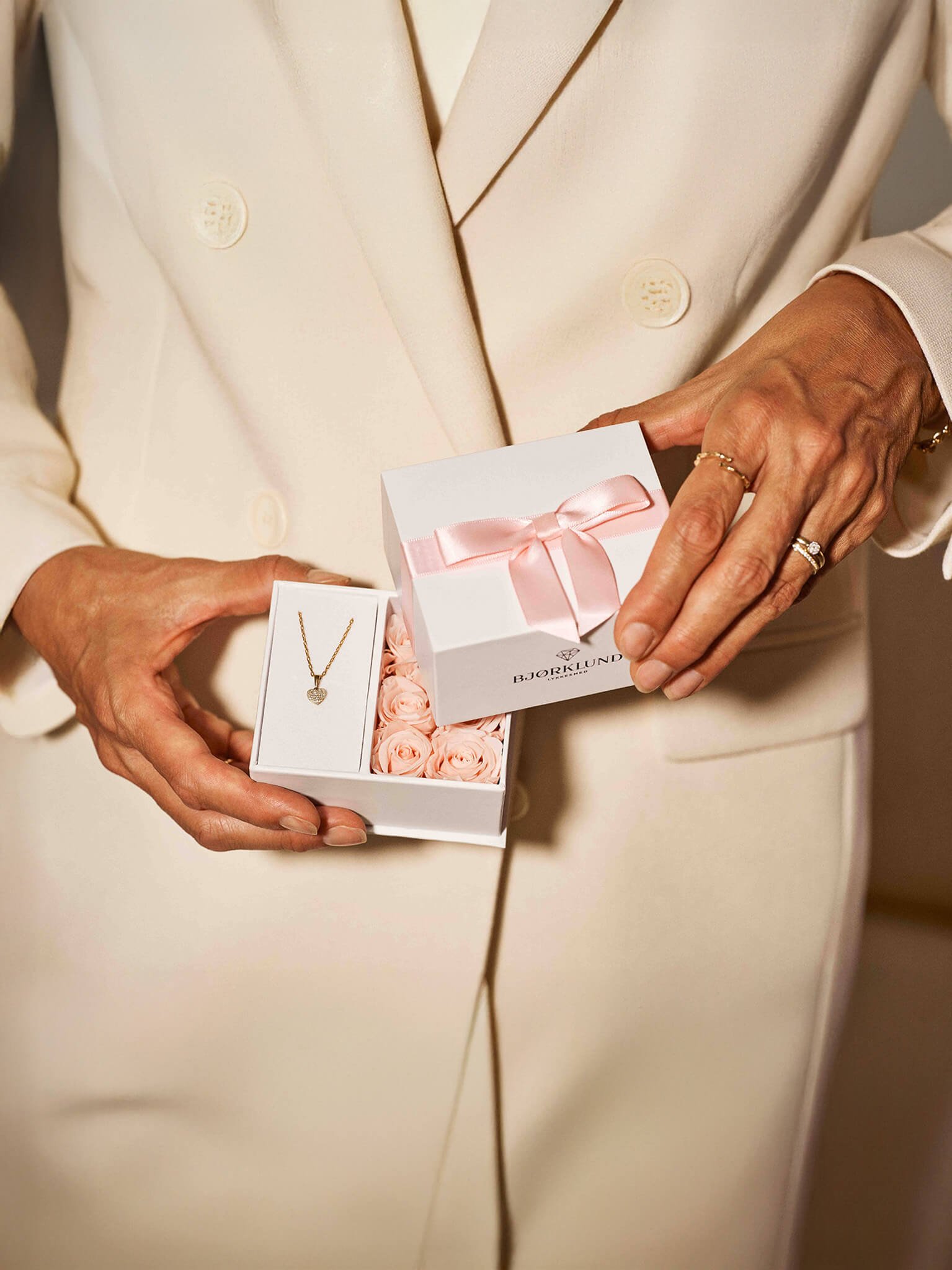 Kvinne i lys blazer holder gaveeske fra Bjørklund med gullkjede og rosa blomster.