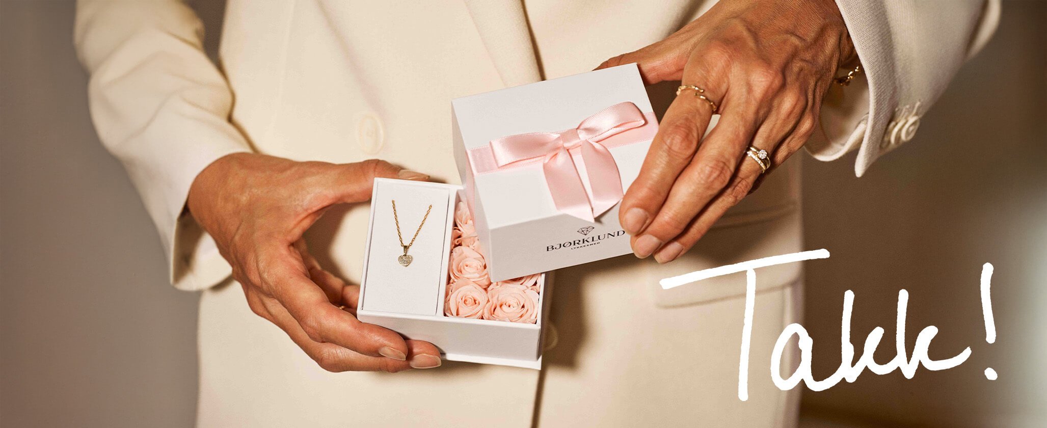 Gavekonseptet vårt! Kvinne i lys blazer holder Bjørklund-gaveeske med gullhjerteanheng og rosa blomster. Tekst: "Takk!"