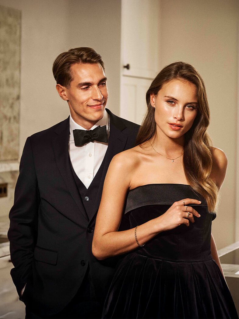 Kvinne i sort kjole og mann i dress står sammen, begge pyntet med smykker og tilbehør.