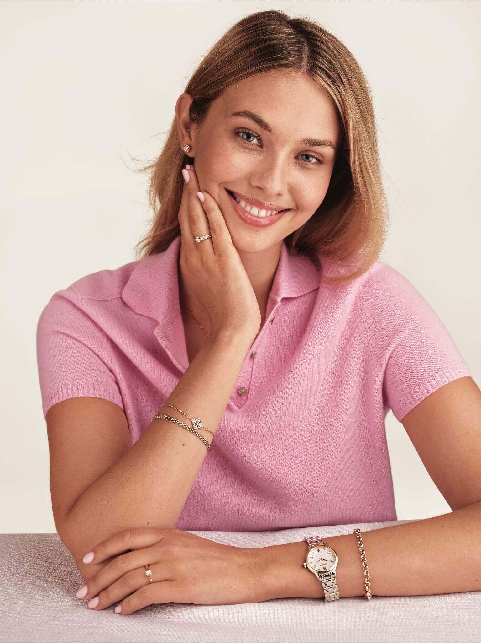 En vakker, ung og blond kvinne med rosa pique, masse forskjellige smykker og klokke, lener ene hånden på haken mens hun smiler til kamera. 
