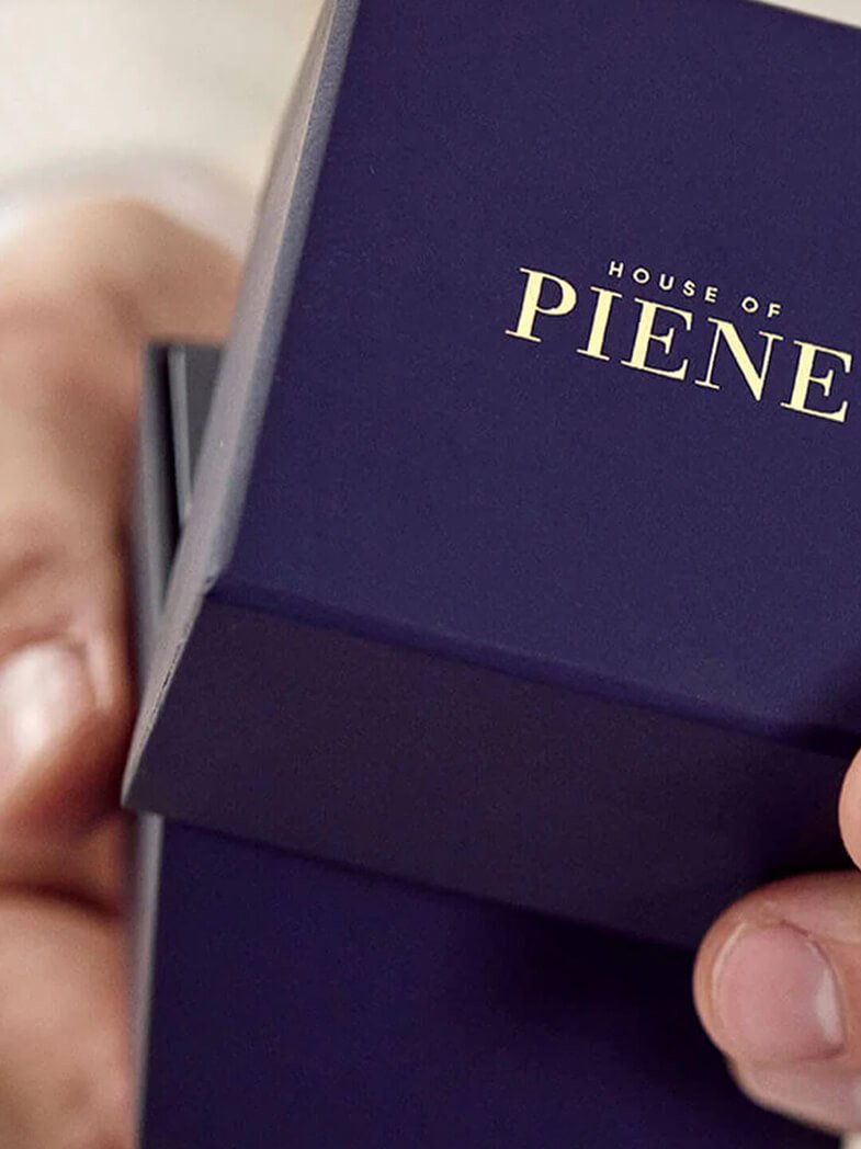 To hender holder mørkeblå smykkeeske med gulltrykk 'House of Piene'. Esken vises delvis åpen med fokus på merkevarelogo og premium finish.