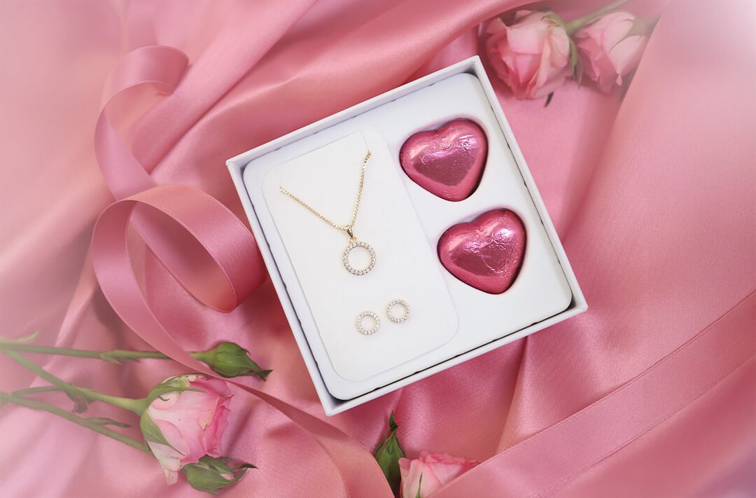 Kjøp en gaveeske til din kjære Smykkesett med gullkjede og øredobber i gaveeske sammen med to rosa sjokoladehjerter på rosa silkestoff.